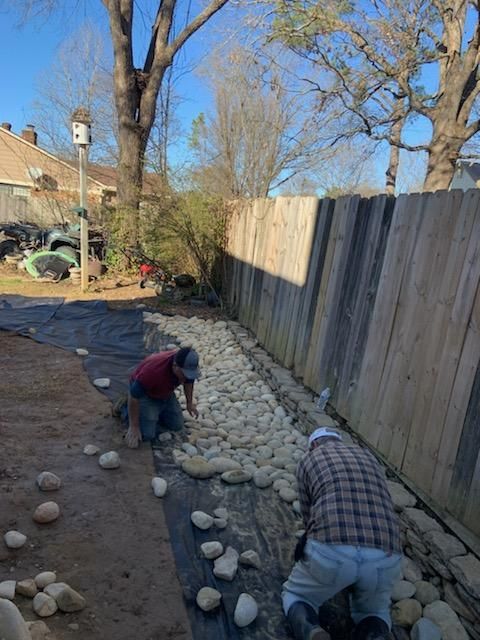 Two workers laying rocks in a gravel trench next to a wooden fence.