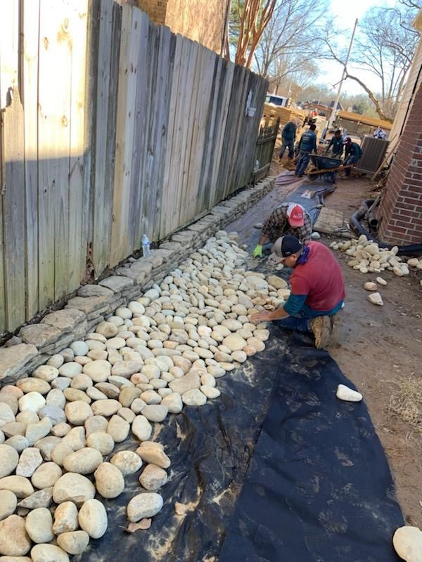 Workers laying river rocks on landscaping fabric near a wooden fence.