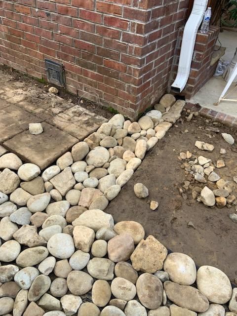 Rocks border a brick wall and walkway, with a downspout emptying onto dirt.
