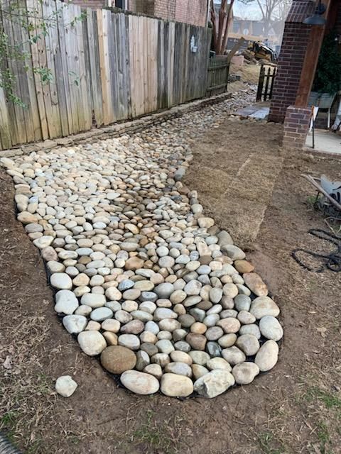 Pathway of smooth, light-colored stones curves through a yard with a wooden fence backdrop.