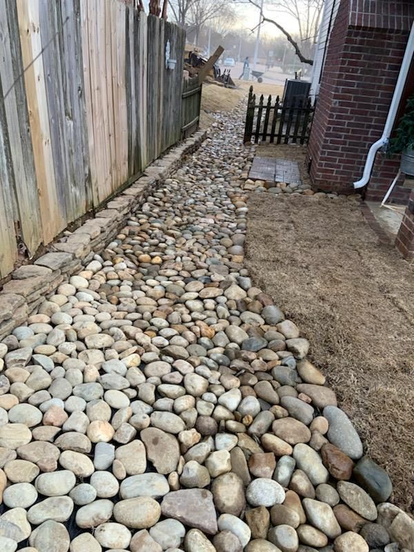 A rock-filled path between a wooden fence and a brick house. Dry grass borders the rocks.