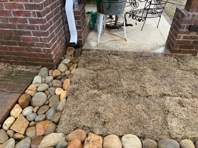 A brick entrance with river rocks and mulch in front of a porch with a white bucket.