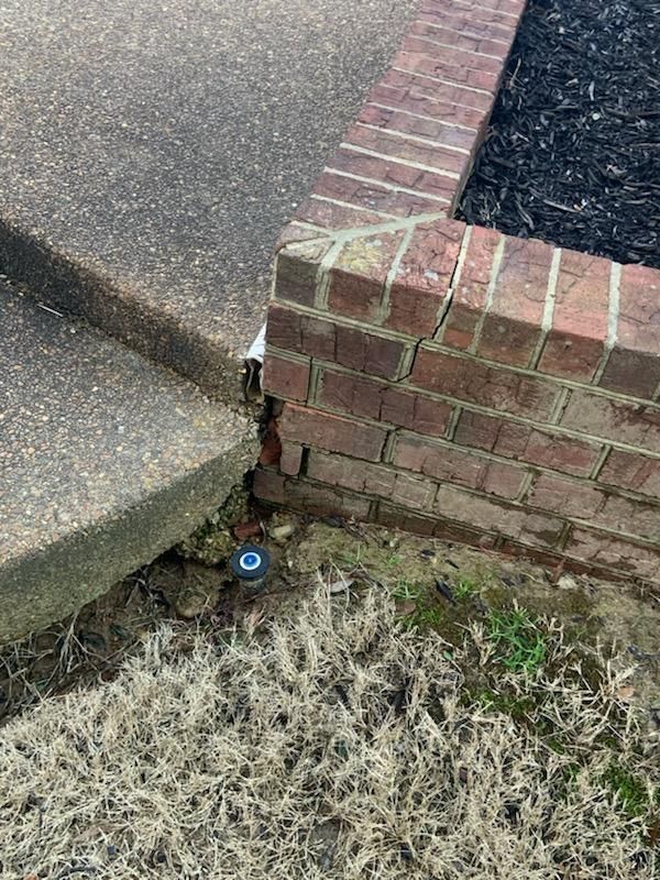 Brick retaining wall separating a concrete walkway and a landscaped area, with a gap where the wall meets the concrete.