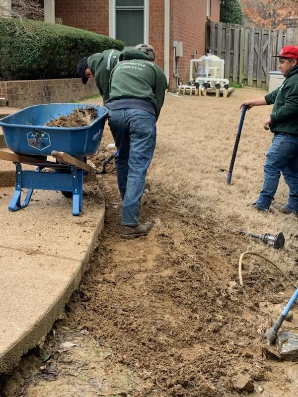 Three landscapers working in a yard. Two dig, one with a wheelbarrow; setting: sunny day.