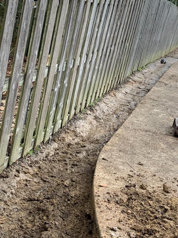 Trench along a weathered wooden fence and concrete border filled with dirt.