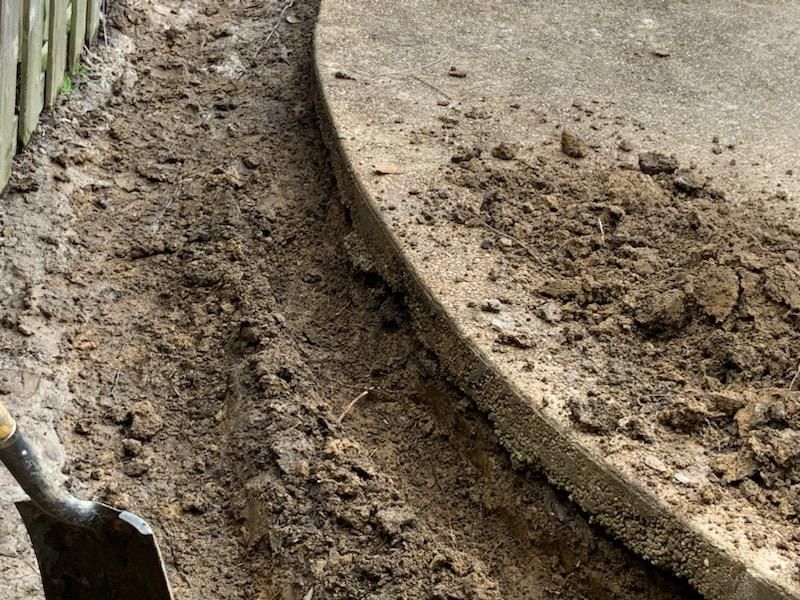 Trench dug next to a curved concrete edge. Soil and a shovel are visible.