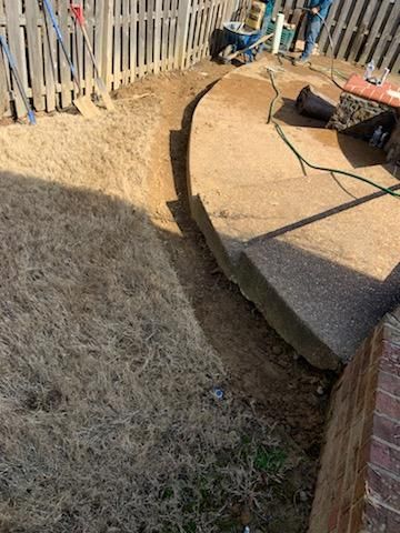 A trench dug along a concrete patio, near a fence and brick wall, with tools and a person in the background.