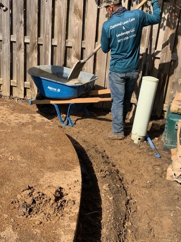 Man shovels cement from a wheelbarrow into a trench in a yard, next to a fence.