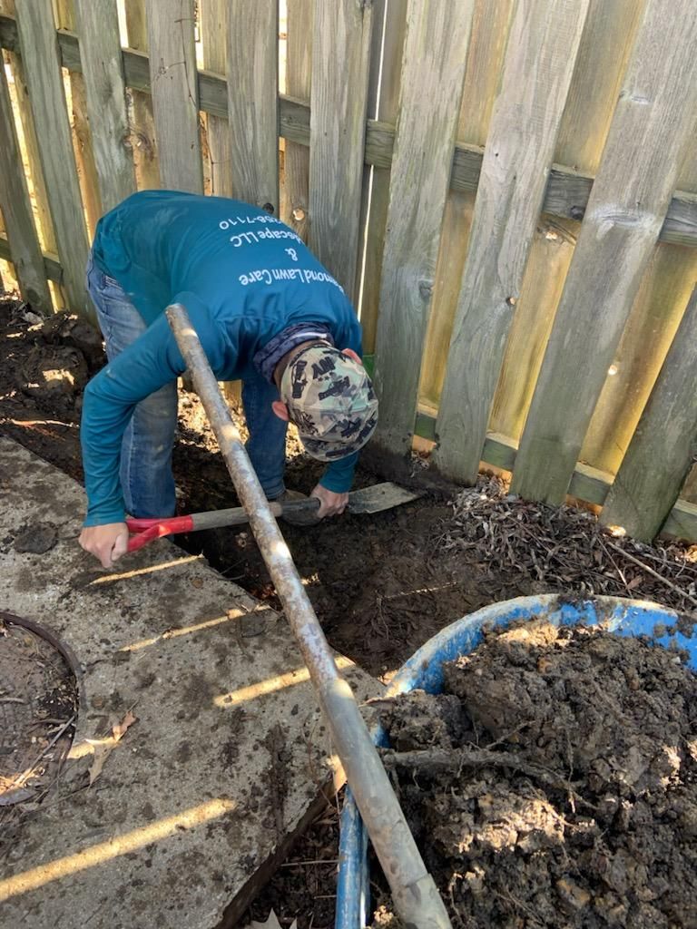 Person digging a trench next to a wooden fence; they are wearing a green shirt and a camouflage hat.