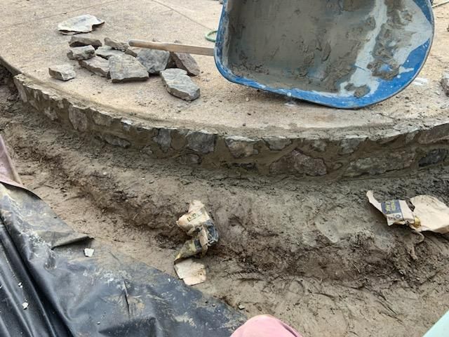 Construction site: A circular stone border with a trench dug around it. A wheelbarrow of cement is nearby.