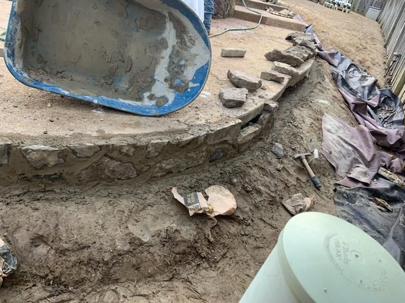 Construction of stone retaining wall and steps.  Wheelbarrow, dirt, hammer, and brush are present.
