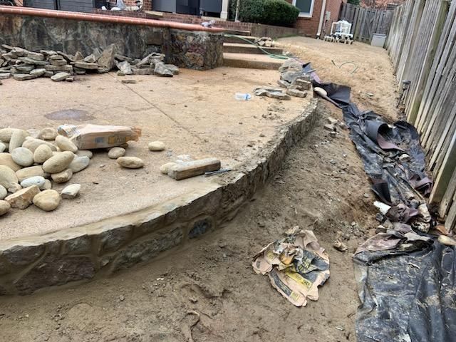 Construction site with stone edging, sand, rocks, and black tarp near a wooden fence.