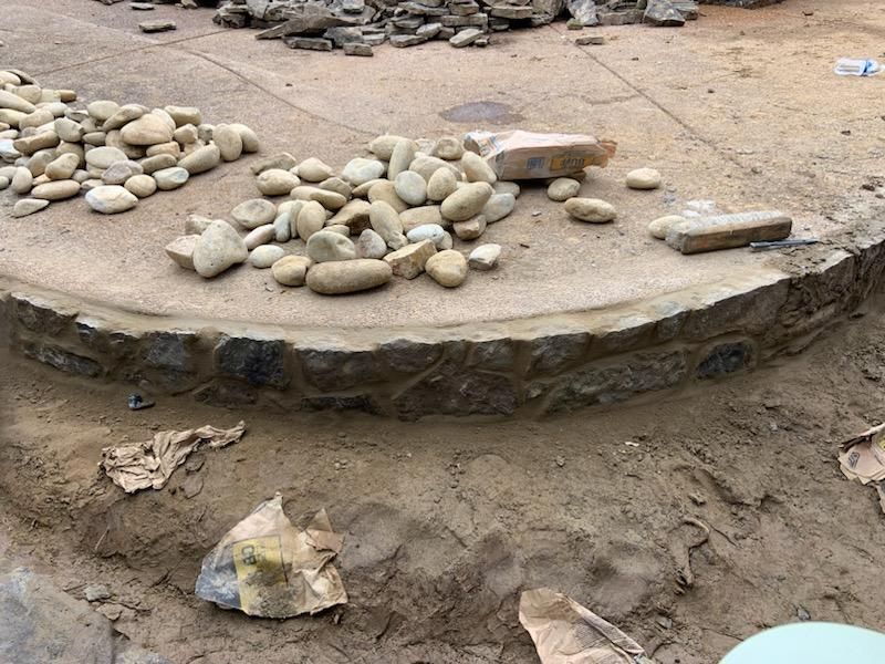 Circular stone retaining wall under construction, with rocks and sand.