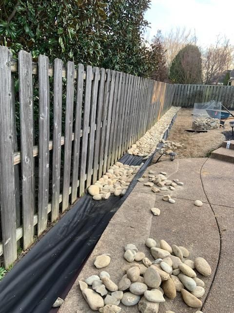 Gravel landscaping along a wooden fence, with black fabric beneath and scattered rocks on the concrete patio.