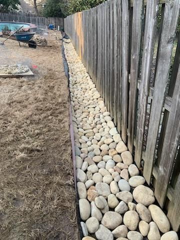 Stone-filled border along a wooden fence in a backyard, with dry grass.