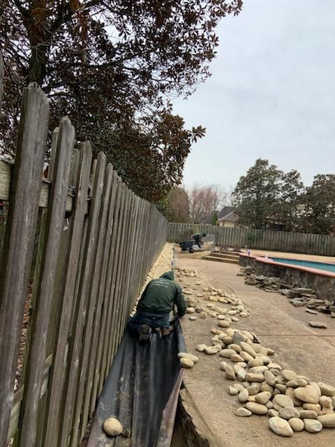 Workers placing river rocks along a concrete patio next to a wooden fence and a pool. Overcast day.