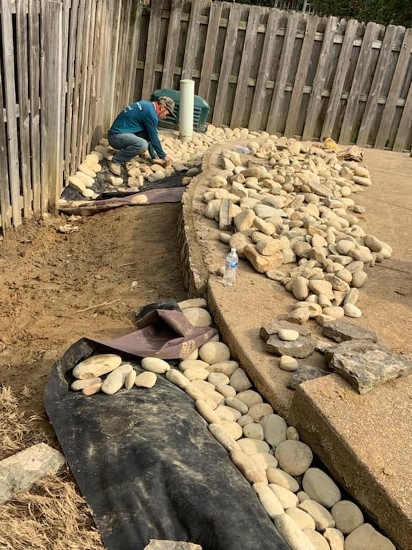 Person landscaping a yard, placing rocks along a black fabric border next to a cement patio and wooden fence.