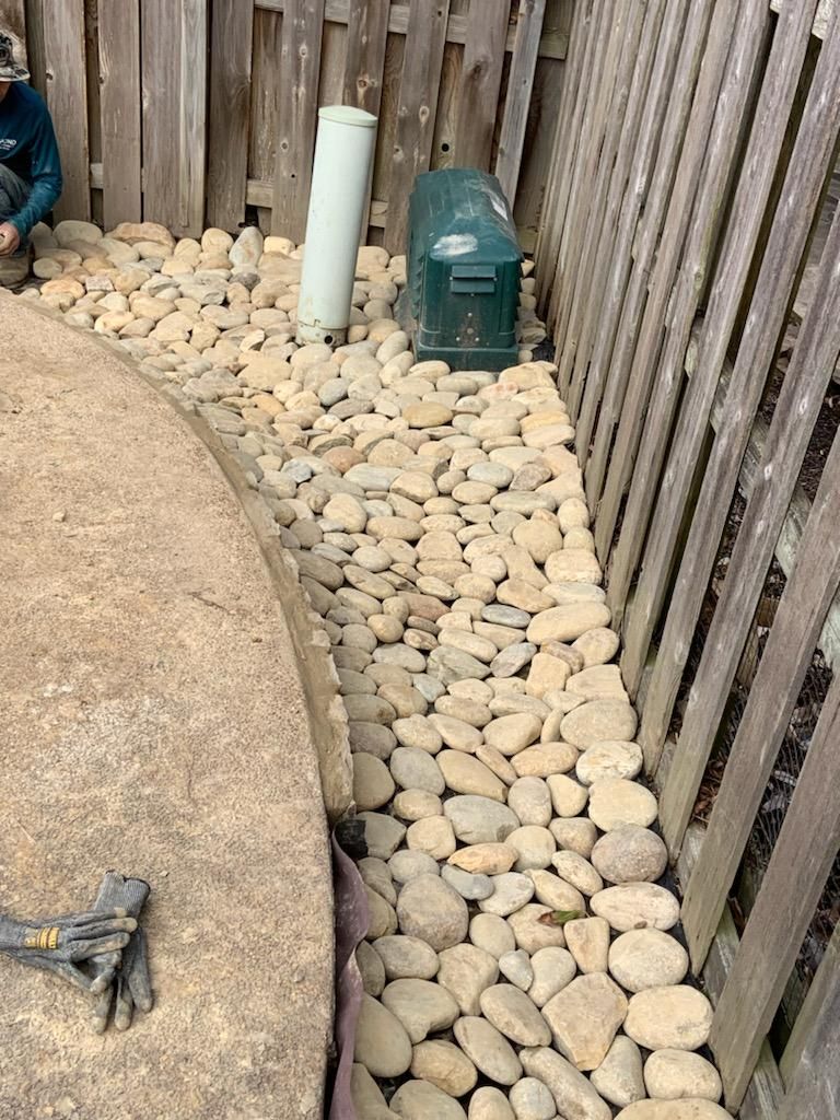 Rocks surround a concrete patio edge and a wooden fence. Green electrical box and white pipe visible.