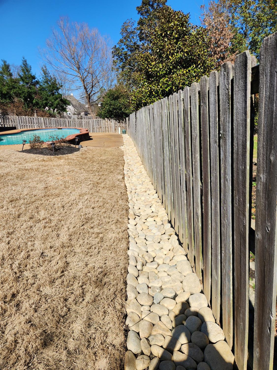 Backyard with a wooden fence, dry grass, and a rock border along the fence.