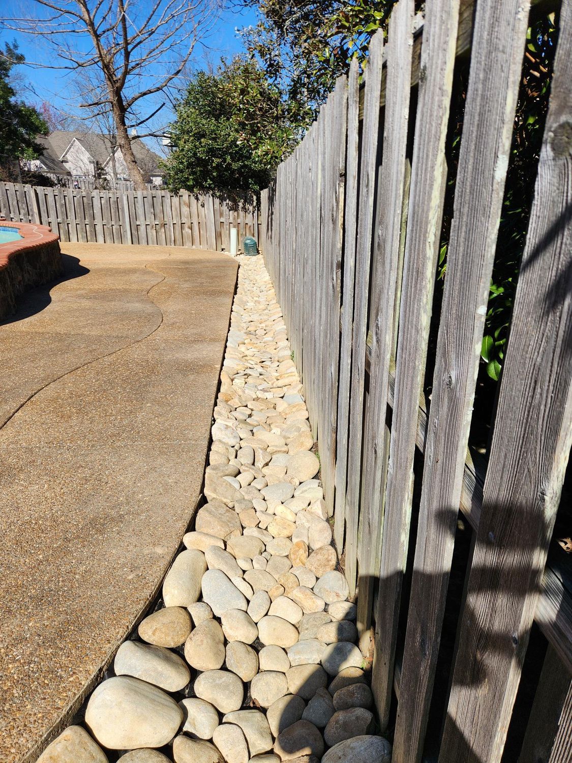 Stonewall rock border beside a wooden fence, alongside a concrete patio. Bright day, outdoor setting.