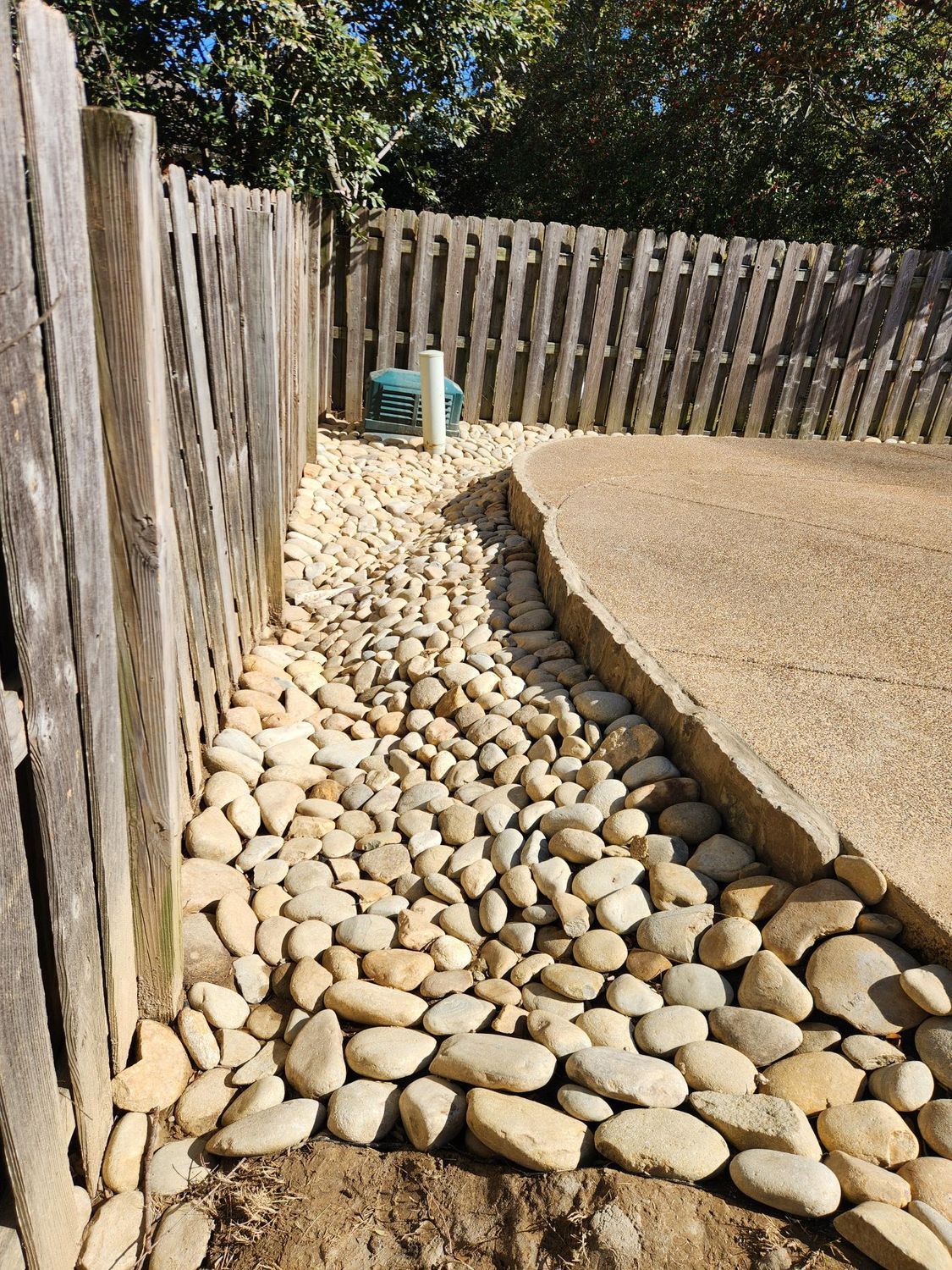 A rock-filled drainage area next to a wooden fence and concrete patio.