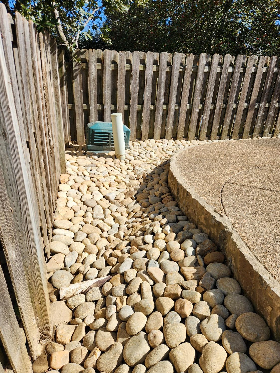 A gravel pathway between a wooden fence and a concrete patio, with a water pipe visible.