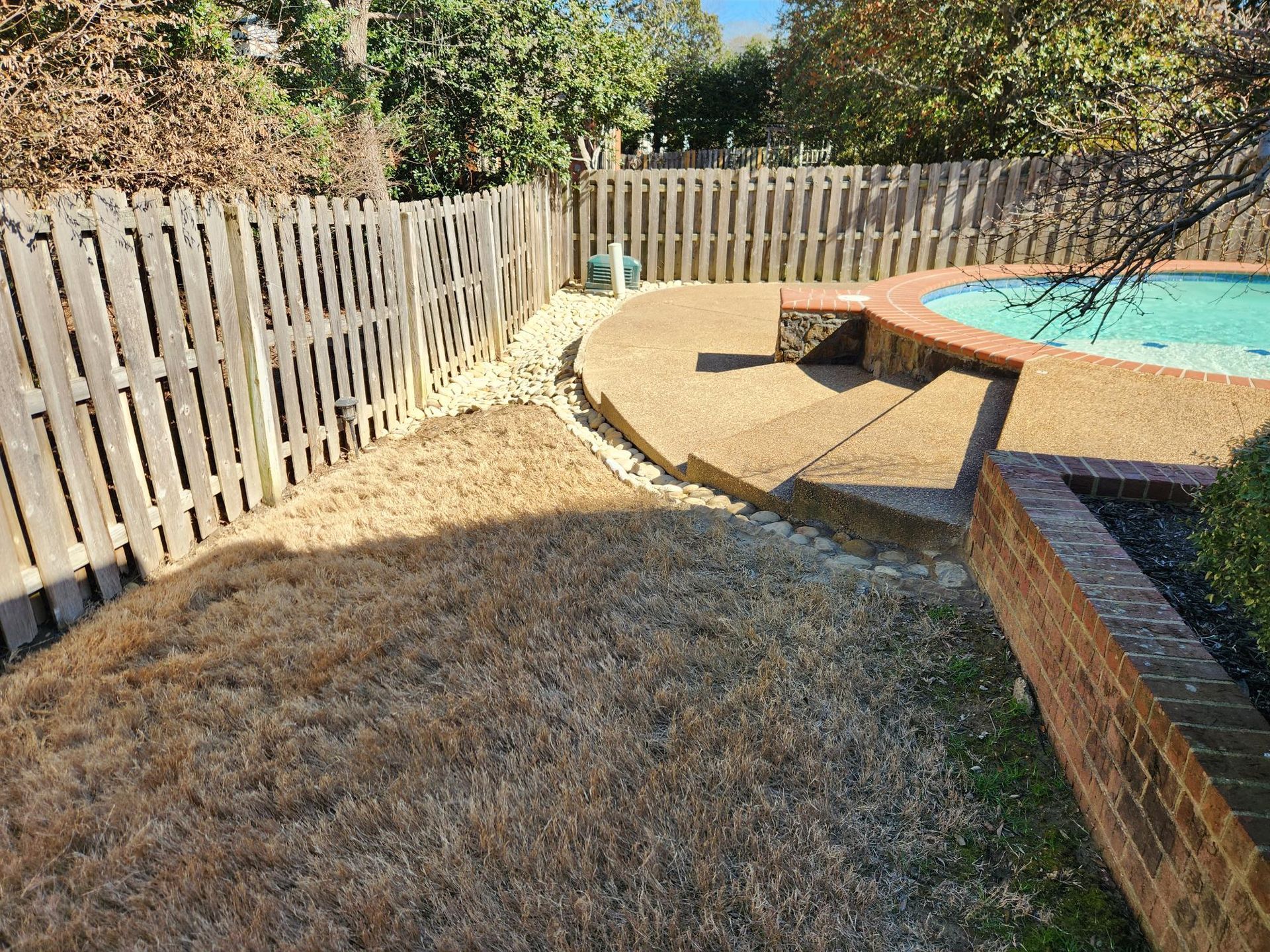 A backyard with a pool and wooden fence. Brown grass and gravel border the pool deck.