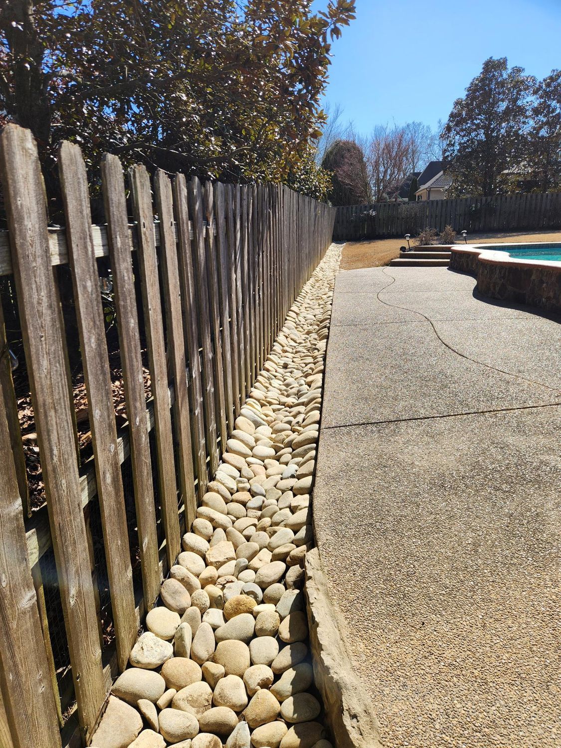 A pebble-filled border alongside a wooden fence and a concrete patio, near a swimming pool.