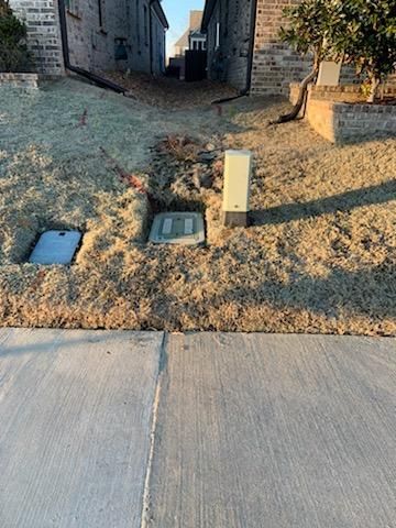 Three utility boxes in a grassy yard next to a sidewalk, with houses in the background.