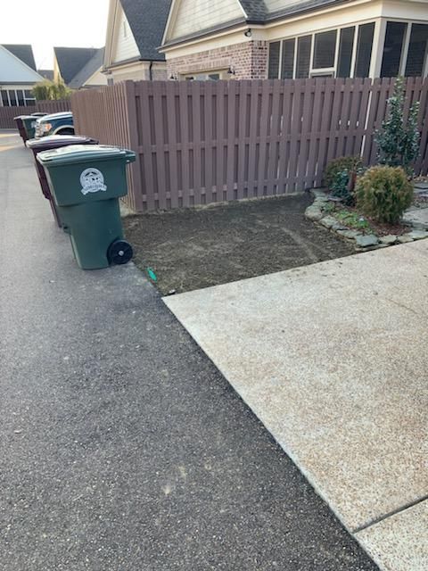 Green trash bins beside a driveway, leading to a brown fence, in front of a house.