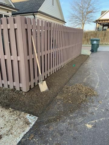 A brown wooden fence with a shovel leaning against it, alongside a freshly dug garden bed.