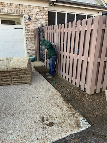 Person in green hoodie working on ground next to brown fence. Stack of sod and garage door visible.