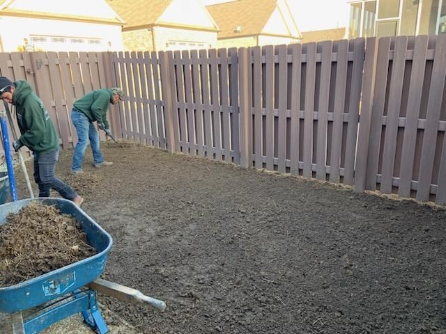 Two people raking dirt in a yard near a brown fence, with a blue wheelbarrow filled with soil in the foreground.
