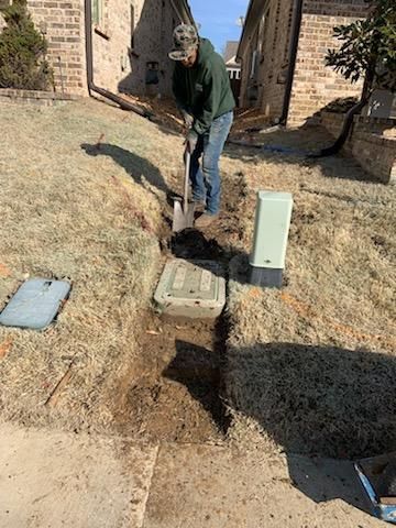 Man digging a trench on a grassy slope near brown brick buildings.