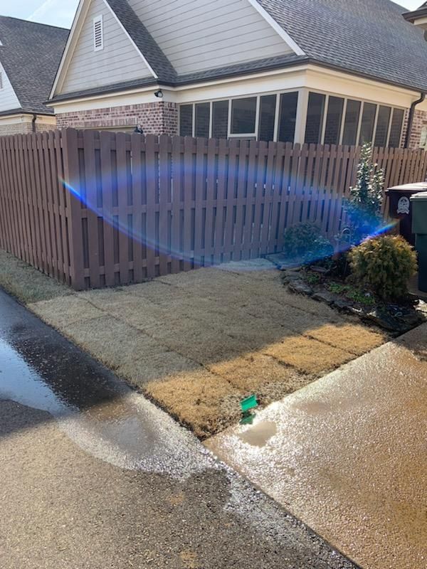 Brown picket fence borders a yard with tan grass, a small bush, and part of a house. Water from a sprinkler.
