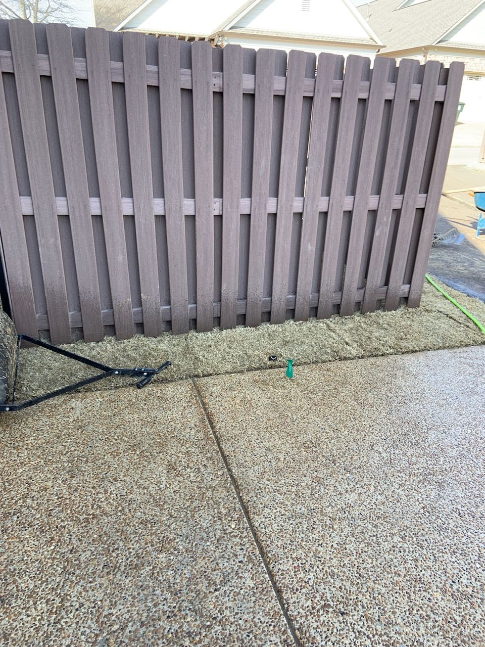Brown wooden fence. A sprinkler is on the grass. Concrete in the foreground.