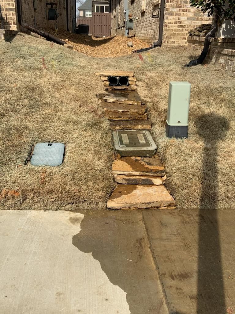 Stone walkway over a water drainage system in a grassy area, with a utility box and building in the background.