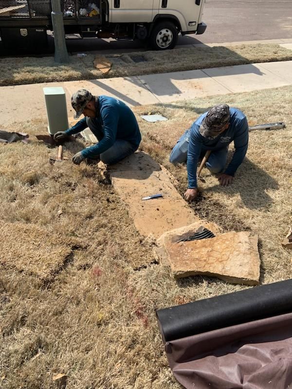 Two men install a stone walkway in dry grass. One kneels by an electrical box, the other holds a tool.