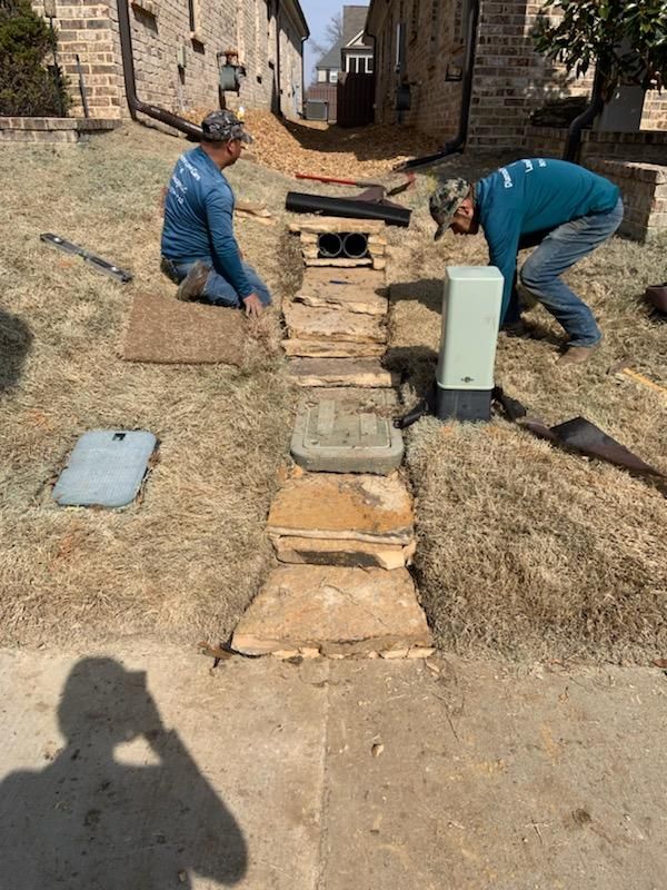 Two workers installing a drainage system in front of a brick house on a sunny day.