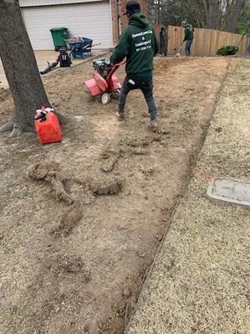 Man using a tiller to prepare a garden bed next to a lawn; other workers and a tree in the background.