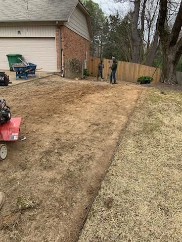 Two people work in a dirt yard near a garage and fence. A red tiller and green trash bin sit nearby.