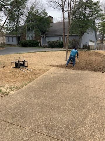 A person mulches a yard near a driveway and a gray house on a cloudy day.