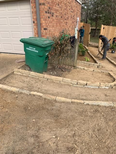 Two people landscaping a yard with stone borders and dirt beds near a green trash can and garage.