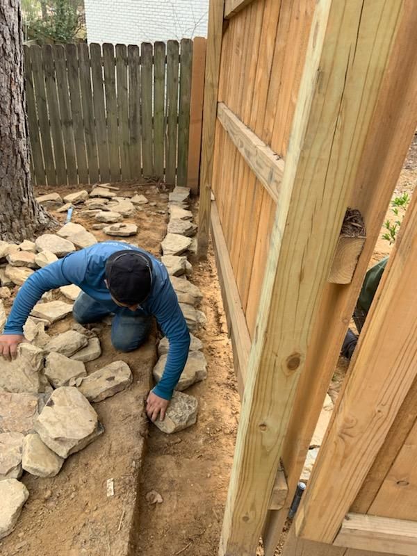 A man is kneeling down in the dirt next to a wooden fence.