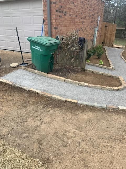 A gravel pathway lined with tan stones curves past a green trash can beside a garage.