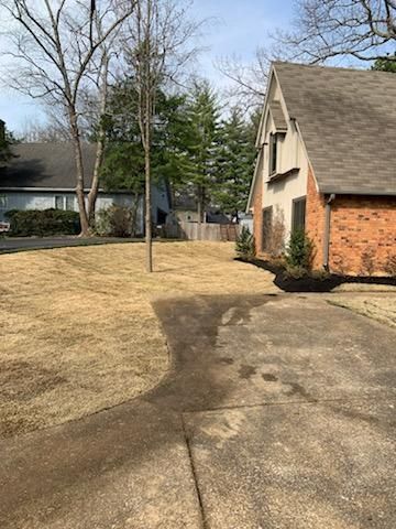 A brown, grassy yard with a concrete driveway leading to a brick house with a brown roof.