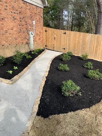 A pathway lined with plants and black mulch leads to a wooden gate near a brick building.