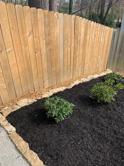 Wooden fence with a bed of dark mulch, stone border, and green shrubs.