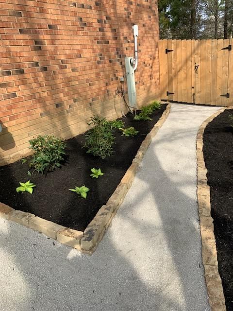 A concrete pathway with a mulch-covered flowerbed beside a brick wall and wooden fence.