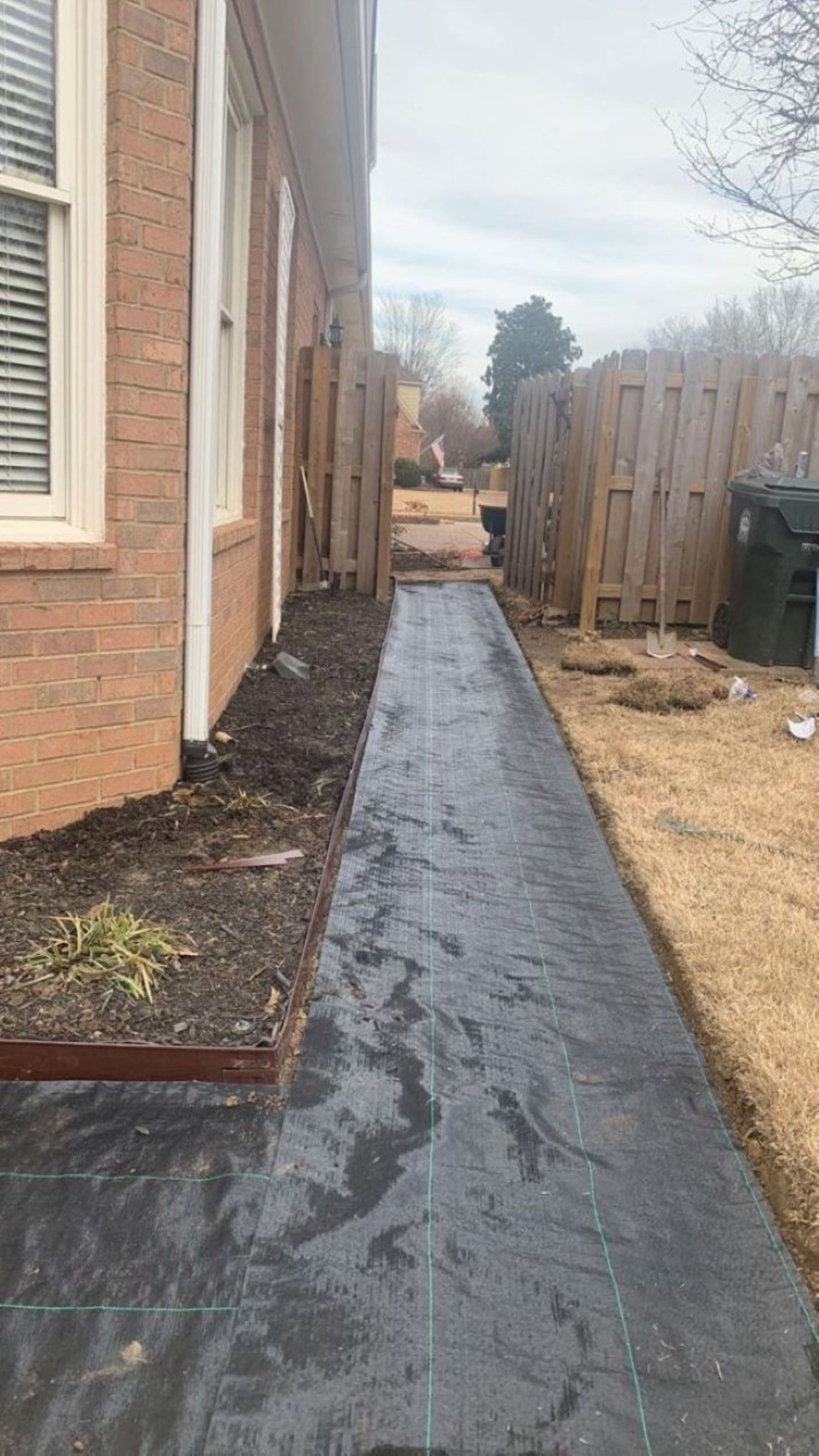 Black fabric walkway between a brick building and a wooden fence, overcast day.